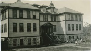 Group of Indigenous youth are standing on grounds and on stairs of Alberni Residential School building.
