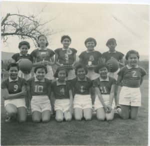 Group of Indigenous youth dressed in athlethic uniforms are posed outdoors, six kneeling and five standing behind them. Two standing are holding basketballs and three are holding trophies.