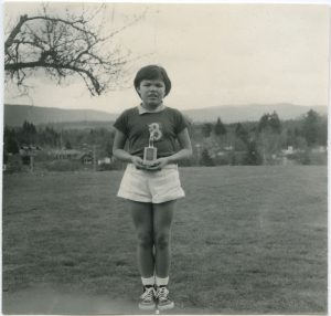 An Indigenous youth is standing in the lawn, dressed in team uniform, and holding a trophy.