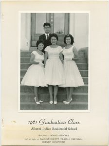 Image of four Indigenous youth dressed in formal clothing, three in white dresses and one in suit and tie, are standing in front of doors of Alberni Residential School. Caption in bottom is listed in description.