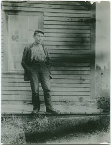Indigenous man with hands in his pant pockets is standing on wooden planks in front of wooden building.