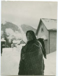 Elderly Indigenous woman wearing a plaid shaw and head scarf is carrying her grandchild on her back in the snow. Buildings are in the background.